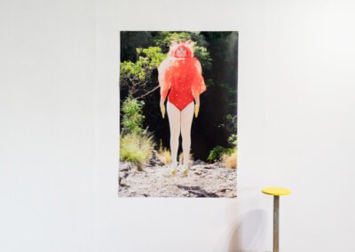 a0 sized photo of a girl suspended mid air in a red mushroom costume in nature. The photo is hang with magnets on a white gallery wall, beneath is a wooden stand with a wooden mushroom sculpture, photo and a smoke alarm