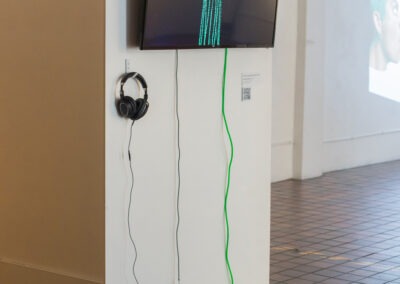 Shot of a tv screen hang on a white wall in a gallery. The TV has a neon cord and black head phones coming out from below