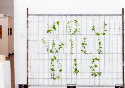 a temporary mesh fence, against a white gallery wall. On the fence is spelled out in pothos vine "you will die"