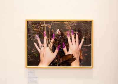 A photo taken of a framed photo on a white wall. The framed photo is of two hands wearing fake nails with the word "cyber break" spelt out across the nails. The hands sit on top of a woody forest floor.