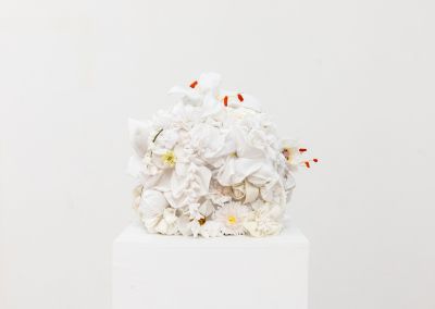 white handmade fabric flowers, appear on top of a plinth within a white walled gallery. The flowers are constructed into a cube.