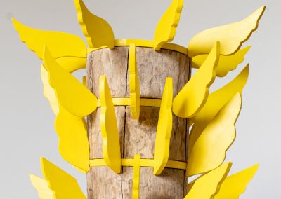 top half of a sculpture made of a found wooden stump and then layers of bright yellow ply. Those layers have bright yellow ply wings extended outwards