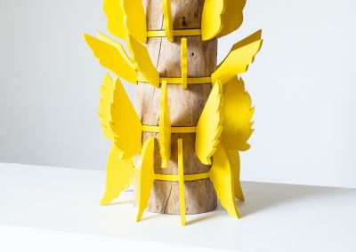 bottom half of a sculpture made of a found wooden stump and then layers of bright yellow ply. Those layers have bright yellow ply wings extended outwards