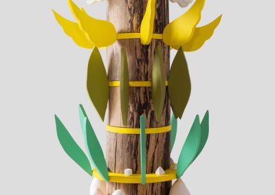 a shot of a wooden sculpture that uses the base of found tree stump. It has 6 rings of bright yellow ply and radiating outwards from those rings are various things per layer. The first layer of ceramic moths and mushrooms, white eggs and pink tulips and a white waterbird. The next layer from the bottom has aqua coloured ply leaves and small unglazed ceramic shapes. The top layer has white ply angel wings. The layer below has bright yellow angel ply wings and the next has olive green ply leaves