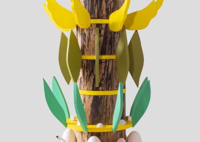 a shot of a wooden sculpture that uses the base of found tree stump. It has 6 rings of bright yellow ply and radiating outwards from those rings are various things per layer. The first layer of ceramic moths and mushrooms, white eggs and pink tulips and a white waterbird. The next layer from the bottom has aqua coloured ply leaves and small unglazed ceramic shapes. The top layer has white ply angel wings. The layer below has bright yellow angel ply wings and the next has olive green ply leaves