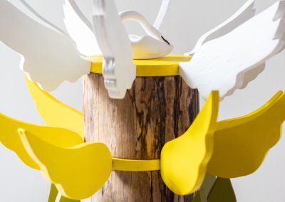 the top half of a sculpture made out of salavged wood log, the very top lies a white ceramic swan, surrounded by white plywood wings. Below that there are bright yellow plywood wings and below that are olive and aqua plywood leaves
