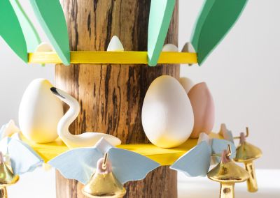 a close up shot of a wooden sculpture that uses the base of found tree stump. This close up shot shows the bottom of the sculpure. we can see 3 bright yellow plywood layers. Radiating outwards from those rings are various things per layer. The first (bottom) layer has ceramic moths and mushrooms, white eggs and pink tulips and a white waterbird. The next layer from the bottom has aqua coloured ply leaves and small unglazed ceramic shapes.
