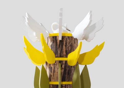 the top half of a sculpture made out of salavged wood log, the very top lies a white ceramic swan, surrounded by white plywood wings. Below that there are bright yellow plywood wings and below that are olive and aqua plywood leaves
