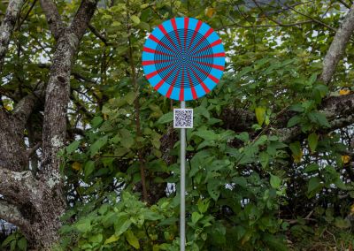 A photo taken outside at Noosa, that shows an art sign post, the post has a round blue psychedelic graphic and sits in front of a tree.