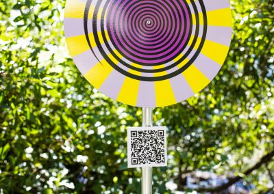 A photo taken outside at Noosa, that shows an art sign post (close up). The post has a round purple, yellow and black psychedelic graphic and sits in a park amongst trees.
