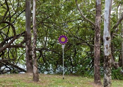 A photo taken outside at Noosa, that shows an art sign post in the middle. The post has a round purple, yellow and black psychedelic graphic and sits in a park amongst trees.