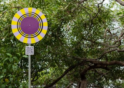 A photo taken outside at Noosa, that shows an art sign post in the middle. The post has a round purple, yellow and black psychedelic graphic and sits in a park in front of trees on the river.
