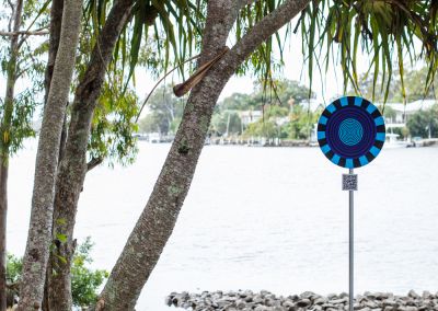 A photo taken outside at Noosa, that shows an art sign post. The post has a round design with a blue and red psychedelic graphic and sits in a park in front of Noosa river.