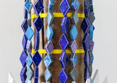 cropped photo of a sculpture shot on a neutral white background. The sculpture is formed using a copper log, that has been sawn into segments. Bright Yellow ply disks appear evenly between the segments, we see 3 in this photo. White ply wings appear at the bottom of the sculpture, we see 2/3rd of these. A rings of light purple ceramic eyes circles the top and hooks appear out of those eyes, with brightly blue glazed diamond shaped ceramics dangle down the length of the sculpture so only glimpses of the original copper log peek through. The diamond ceramics are meant to evoke a stylised waterfall and are in different shades of blue.