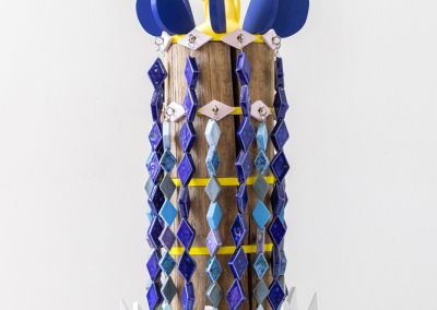 photo of a sculpture shot on a neutral white background. The sculpture is formed using 50 cm of copper log, that has been sawn into 6 segments. Bright Yellow ply disks appear evenly between the segments. Eight ply solid blue wings are attached to the top which circles a stylised yellow star figure in ceramic. White ply wings appear at the bottom of the sculpture. Two rings of light purple ceramic eyes circle the top and hooks appear out of those eyes, with brightly blue glazed diamond shaped ceramics dangle down the length of the sculpture so only glimpses of the original copper log peek through. The diamond ceramics are meant to evoke a stylised waterfall and are in different shades of blue.