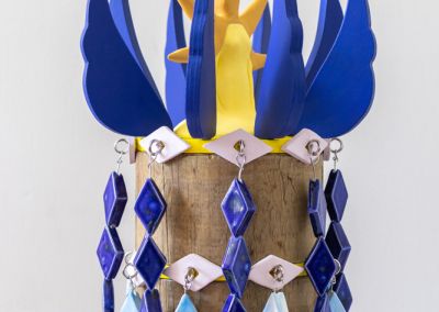 cropped photo of a sculpture shot on a neutral white background. The sculpture is formed using 50 cm of copper log, that has been sawn into 6 segments. Bright Yellow ply disks appear evenly between the segments, we see the top 3 in this photo. Eight ply solid blue wings are attached to the top which circles a stylised yellow star figure in ceramic. Two rings of light purple ceramic eyes circle the top and hooks appear out of those eyes, with brightly blue glazed diamond shaped ceramics dangle down the length of the sculpture so only glimpses of the original copper log peek through. The diamond ceramics are meant to evoke a stylised waterfall and are in different shades of blue.