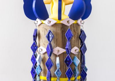 cropped photo of a sculpture shot on a neutral white background. The sculpture is formed using 50 cm of copper log, that has been sawn into 6 segments. Bright Yellow ply disks appear evenly between the segments, we see the top 3 in this photo. Eight ply solid blue wings are attached to the top which circles a stylised yellow star figure in ceramic. Two rings of light purple ceramic eyes circle the top and hooks appear out of those eyes, with brightly blue glazed diamond shaped ceramics dangle down the length of the sculpture so only glimpses of the original copper log peek through. The diamond ceramics are meant to evoke a stylised waterfall and are in different shades of blue.