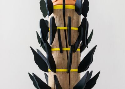 a sculpture shot on a neutral white background. The sculpture is made out of a 50cm tall cylindrical wooden stump (a copper log). The stump is evenly cut into six segments with yellow painted ply wedged between the segments. Radiating outward from each yellow ply ring are forest green painted plywood wings. The wings gradually get smaller as they reach the top in attempt to evoke a pine tree shape. At the top of the sculpture is a terracotta cone with 2 moth shapes. On top of the terracotta cone rests a porcelain tulip flower which has been painted in pastel pink.