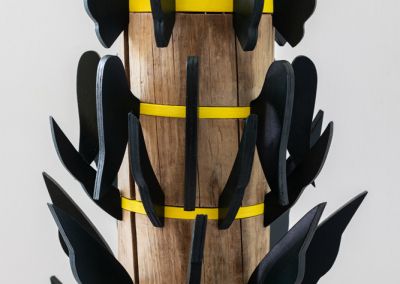 a cropped photo of a sculpture shot on a neutral white background. The sculpture is made out of a 50cm tall cylindrical wooden stump (a copper log). The stump is evenly cut into six segments with yellow painted ply (we see five in this photo) wedged between the segments. Radiating outward from each yellow ply ring are forest green painted plywood wings. The wings gradually get smaller as they reach the top in attempt to evoke a pine tree shape. At the top of the sculpture is a terracotta cone with 2 moth shapes On top of the terracotta cone rests a porcelain tulip flower which has been painted in pastel pink.