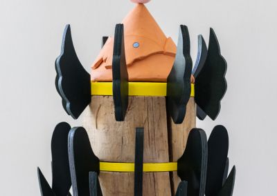 a cropped photo of a sculpture shot on a neutral white background. The sculpture is made out of a 50cm tall cylindrical wooden stump (a copper log). The stump is evenly cut into six segments with yellow painted ply (we see three in this photo) wedged between the segments. Radiating outward from each yellow ply ring are forest green painted plywood wings. The wings gradually get smaller as they reach the top in attempt to evoke a pine tree shape. At the top of the sculpture is a terracotta cone with 2 moth shapes On top of the terracotta cone rests a porcelain tulip flower which has been painted in pastel pink.