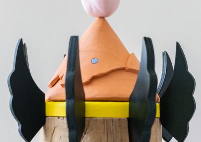 a cropped photo of a sculpture shot on a neutral white background. The sculpture is made out of a 50cm tall cylindrical wooden stump (a copper log). The stump is evenly cut into six segments with yellow painted ply (we see two in this photo) wedged between the segments. Radiating outward from each yellow ply ring are forest green painted plywood wings. The wings gradually get smaller as they reach the top in attempt to evoke a pine tree shape. At the top of the sculpture is a terracotta cone with 2 moth shapes On top of the terracotta cone rests a porcelain tulip flower which has been painted in pastel pink.