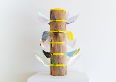 a sculpture made of a found wooden stump and then 6 layers of bright yellow pine. Those layers have iridescent stained glass wings extended outwards in different colours. Each level has different shapes wings.
