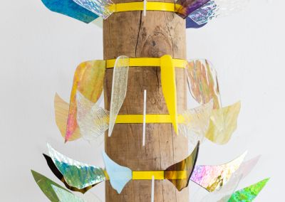 a sculpture made of a found wooden stump and then 6 layers of bright yellow pine. Those layers have iridescent stained glass wings extended outwards in different colours. Each level has different shapes wings.