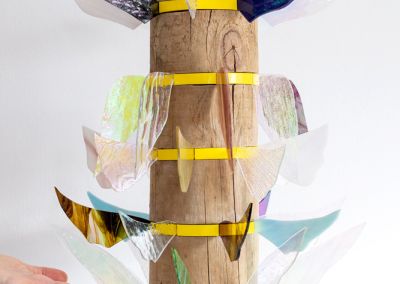 a sculpture made of a found wooden stump and then 6 layers of bright yellow pine. Those layers have iridescent stained glass wings extended outwards in different colours. Each level has different shapes wings. Their is a hand reference size to show that the work is approx 50cm.
