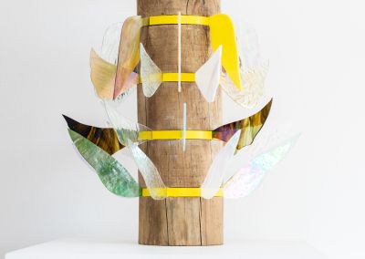 close up of a sculpture made of a found wooden stump with layers of bright yellow pine (in the photo you can see four out of six layers). Those layers have iridescent stained glass wings extended outwards in different colours. Each level has different shapes wings.